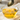 Nahaufnahme und Makroansicht einer kleinen, transparenten Glasschale gefüllt mit klarem, gold-gelbem Agavendicksaft. Ein silberner Löffel hängt über der Schale, von dem ein dicker Strahl der Flüssigkeit zurück in die Schale tropft. Die Schale steht auf einem weißen Tuch auf einer hellen Holzoberfläche.